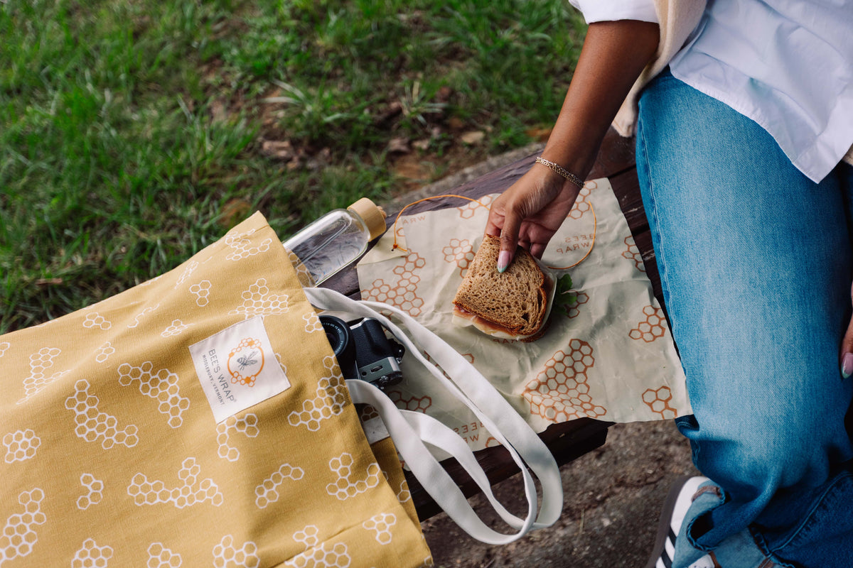 Person holding a sandwich next to a yellow bag with a bee pattern on a grassy background