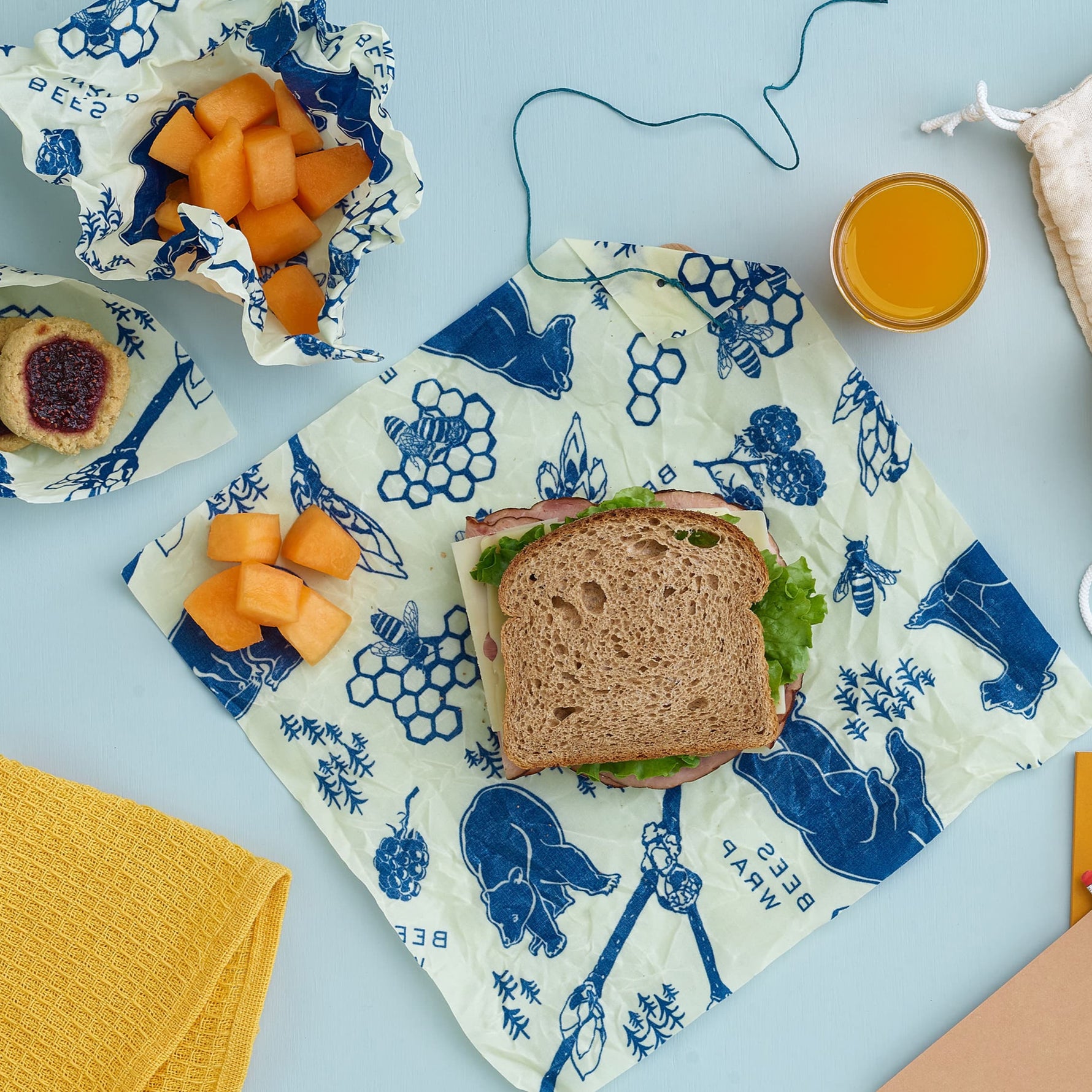 Bees and Bears reusable beeswax wraps with a sandwich, fruit, and juice on a light blue background