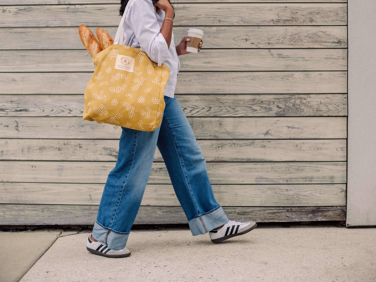 Person walking outdoors with a yellow reusable bag and a coffee cup.