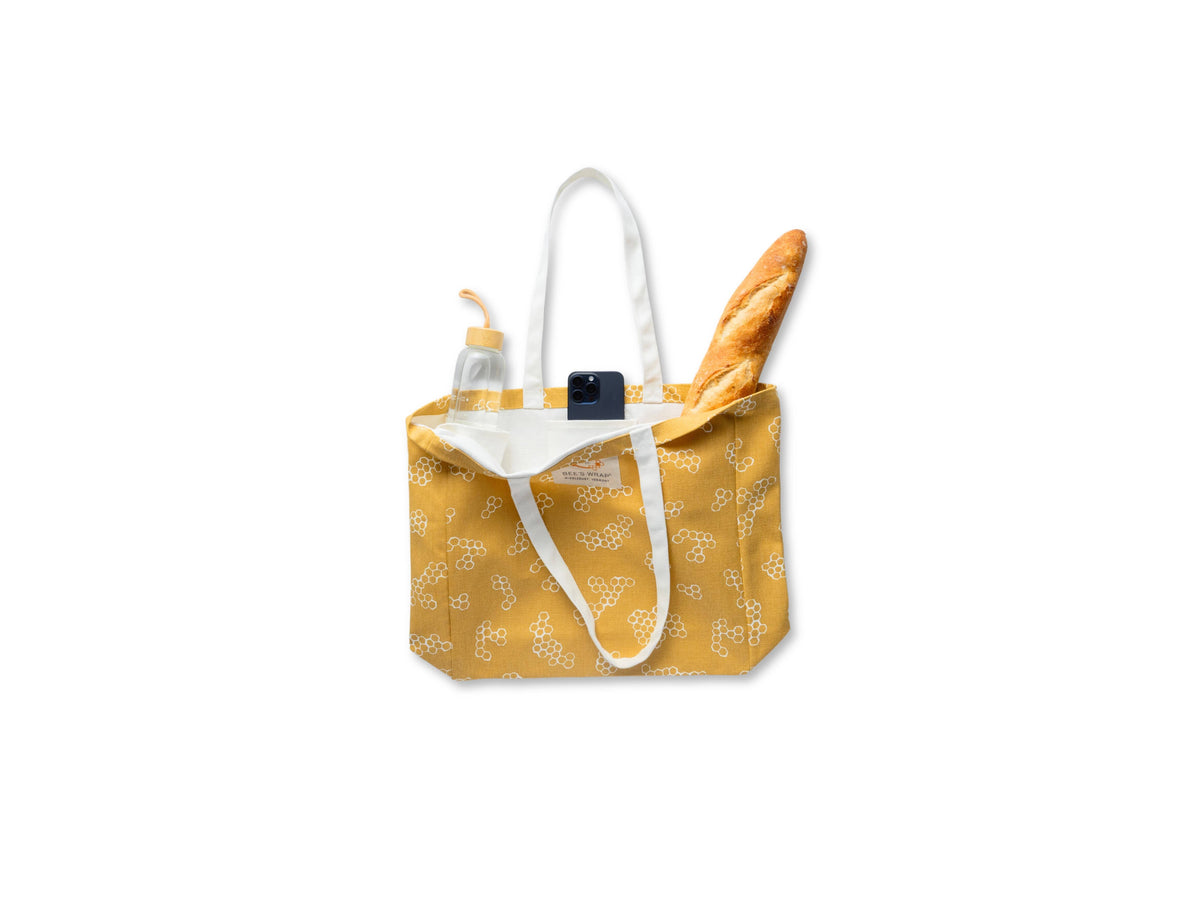 Yellow tote bag with a white strap, containing a bottle and a loaf of bread on a white background